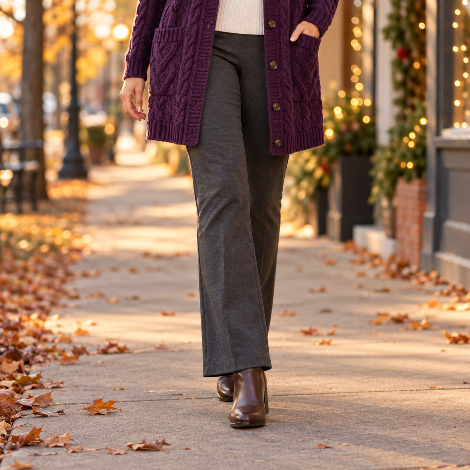 Plum cable-knit cardigan hem over charcoal grey bootcut pants skimming brown ankle boots on a leaf-covered sidewalk